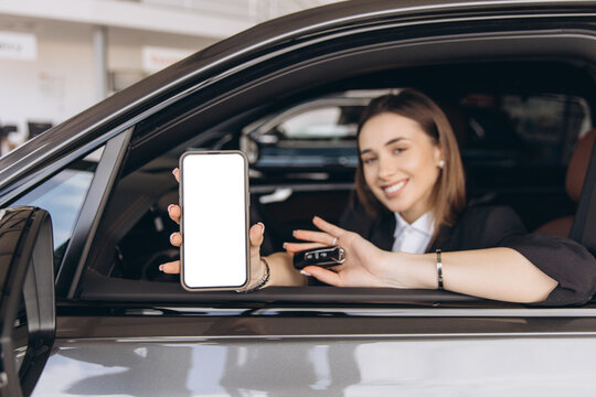 Saleswoman showing smartphone and car key inside new vehicle