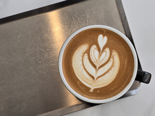 beautifully crafted latte with intricate foam art sits on stainless steel surface, showcasing heart and leaf design. warm tones of coffee create cozy atmosphere