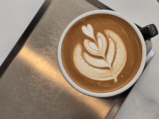 beautifully crafted latte with intricate foam art sits in black cup on metallic surface, showcasing heart and leaf design that adds artistic touch to beverage