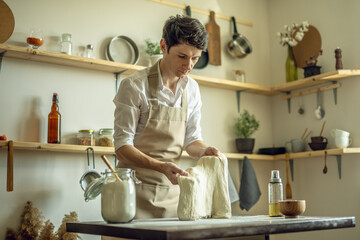A baker in an apron kneads the dough with his hands and sprinkles it with flour to bake delicious homemade bread.