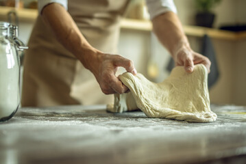 A baker in an apron kneads the dough with his hands and sprinkles it with flour to bake delicious homemade bread.