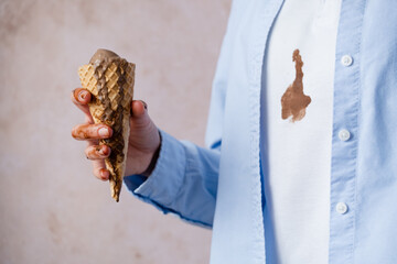 A person holding chocolate ice cream cone, spilled ice cream on white clothes. Ice cream flowing from the cone onto the hand. Concept of daily food stains on clothes