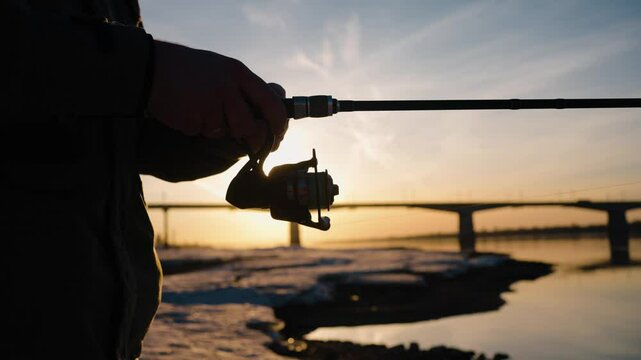 Winter sunset casting golden light on lone fisherman winding fishing line onto spinning rod reel, river bridge reflection mirroring serene landscape