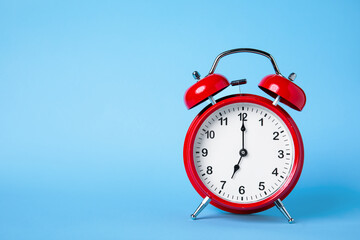 A traditional red alarm clock is displayed against a soft blue background. The clock shows the time near twelve o'clock, symbolizing time management and urgency.
