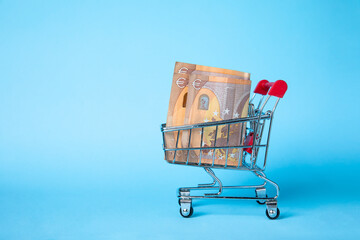 Miniature shopping cart filled with Euro banknotes against a blue background, symbolizing finance,...