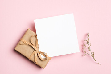 Square white blank card mockup paired with a kraft paper gift box and delicate flower on soft pink...