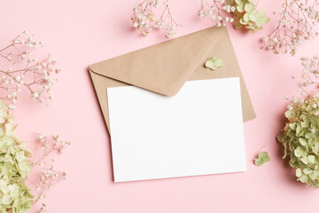 Blank wedding invitation card mockup with craft envelope and trendy flowers decor, top view, white card with copy space on pink