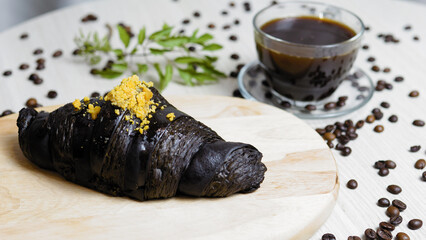 Delicious charcoal croissant with cheese topping lying on wooden board, near cup of coffee and coffee beans scattered on white table, creating inviting breakfast scene