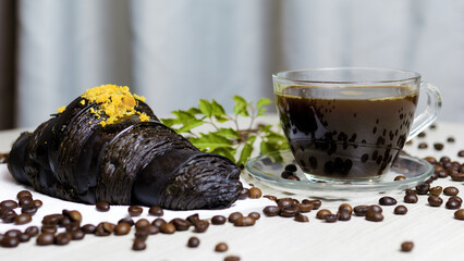 Charcoal croissant with yellow sprinkles and a cup of coffee sitting on a table with scattered coffee beans and a small green branch, creating a delicious and visually appealing breakfast scene