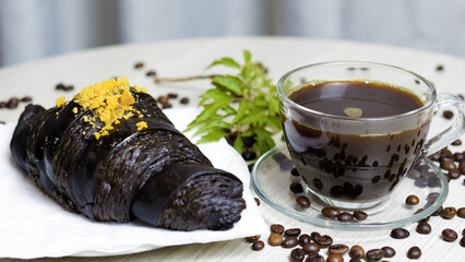 Charcoal croissant with yellow crumbles lying on a white napkin next to a glass cup of hot coffee and coffee beans scattered on a white table, creating a delicious breakfast scene
