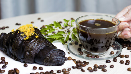 Hand is holding a transparent glass cup of hot black coffee near a black croissant with yellow crumbs and coffee beans scattered on a white table with a green leaf