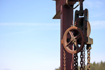 
A rusty metal mechanism with chains and a gear attached to a dark red stand, with a light blue sky...