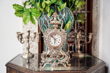 An ornate vintage clock with intricate designs is placed on a glass-topped wooden table. Behind it, a decorative vase with green foliage and two antique candle holders are visible.