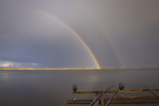 Fishing rods on pier over lake with double rainbow after rain, peaceful natural landscape - Powered by Adobe