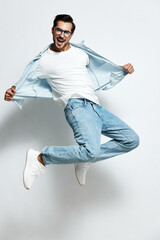 Energetic jumping happy young caucasian man wear white t-shirt and blue jeans with denim jacket isolated on plain light background studio portrait. People lifestyle concept