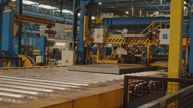 A stack of steel plates on a roller bench are being bound together by a large machine