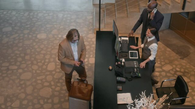High angle view of female receptionist returning passport and back card to guest after check in process at hotel front desk while manager talking on cell phone nearby