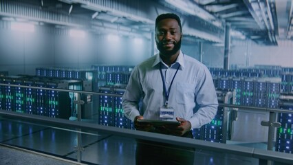 Big Data Center: African American Specialist with Tablet Computer Standing in Storage, Looking at Camera. Server Racks for Cloud Computing Technology. Information Digitalization Concept. Medium Shot.