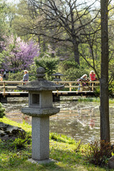 A traditional Japanese stone lantern stands in a serene garden with a pond and cherry blossoms in the background.