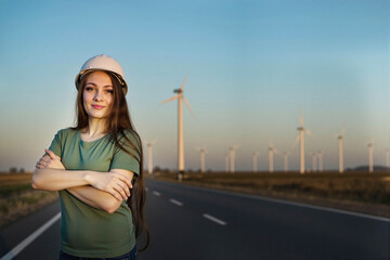 Confident female engineer in hard hat on road at wind farm, arms crossed front view.