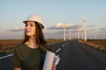 Female engineer in hard hat with plans by curved road at wind farm, looking at turbines view.