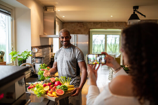 Interracial middle aged couple taking a photo with a smart phone while preparing a healthy dish with fresh fruits and vegetables at home representing a healthy lifestyle and diet