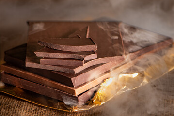 Stack of artisanal dark and milk chocolate bars and pieces, placed on top of each other, surrounded by smoke, close-up.