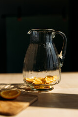 A glass pitcher containing lemon slices, positioned on a wooden surface with sunlight streaming in. Perfect for showcasing homemade lemonade