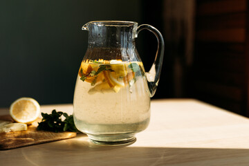 A glass pitcher filled with water, lemon slices, and fresh mint leaves sits on a wooden surface. Sunlight enhances the vibrant colors, capturing the essence of a refreshing homemade lemonade.