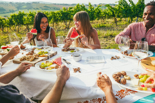 Happy friends having fun drinking white wine and eating watermelon together - Multiracial people doing party at summer time with vineyard in background - Main focus on center blond face