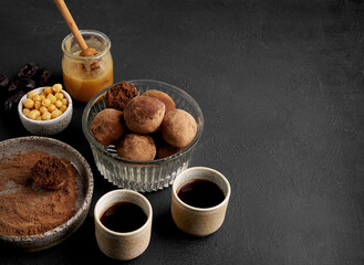 Energy balls of chickpea and dates, sprinkled with cocoa in a glass bowl with cups of coffee nearby on a black background, copy space