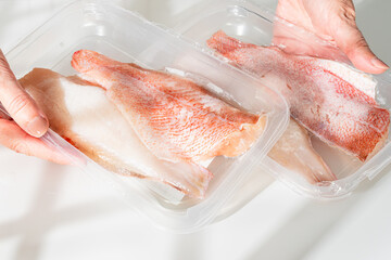  Hands holding two plastic trays of frozen red snapper fillets in bright lighting.