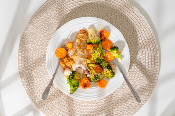 Plate of baked fish fillet served with roasted broccoli and carrots on a white dish.