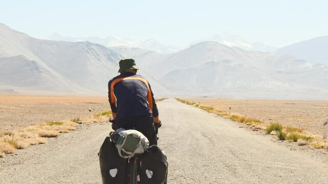 Back view adventurous caucasian male traveller cycle bicycle touring solo on asphalt famous Pamir highway M41 road in remote high Pamir mountains. Pamir highway adventure travel route destination