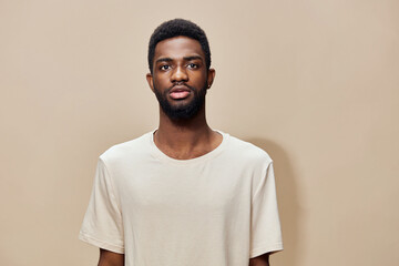 A young Black man with a confident expression wearing a light beige T shirt against a solid light...