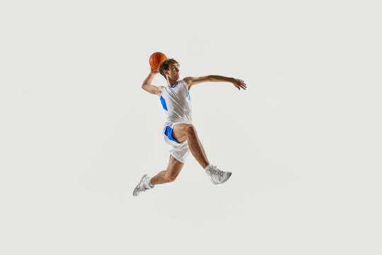 Dynamic photo. Young basketball player mid-jump, leaning forward with focus and grip on ball in power move against white background. Concept of sport, competition, active and healthy lifestyle, game