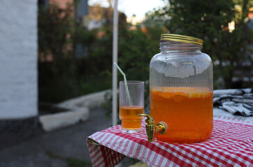 picnic in the village in the yard of the house. glass jar with a tap and an orange refreshing drink on the table and a glass with a straw next to it