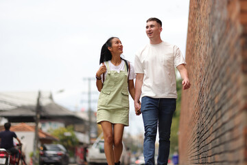 Obraz premium Couple of tourists walking and holding hands on a roadside sidewalk in a tourist attraction area in a city in Thailand. Fellow tourists are walking in a crowded community.