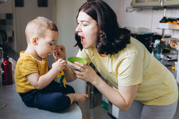 Mother feeding her son with a spoon in the kitchen, sharing a joyful and loving moment of motherhood