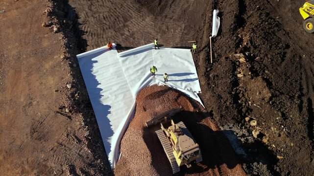 High angle top down late afternoon footage of bulldozer maching covering layers of geotextile with dirt and soil for the construction of a highway. Freeway build site with geosynthetic fabric burried
