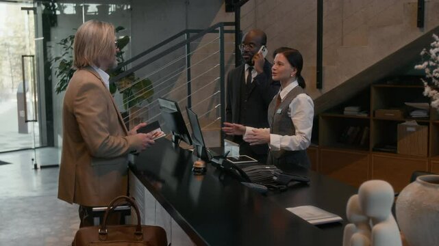 Female hotel receptionist standing at front desk in lobby, returning passport to guest and talking to him while concierge having phone conversation nearby
