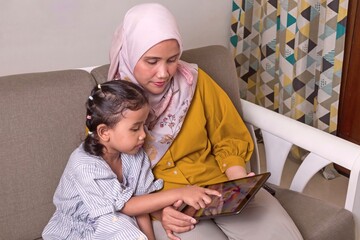 Asian Muslim mother and her little girl relaxing on sofa and playing games together on the tablet pc