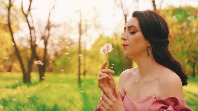 Portrait happy woman holding bouquet ripe dandelions in hands, smiling face enjoys nature. girl blows on dandelions white seeds petals fly, mouth closeup. sun green forest grass trees, park walk fun