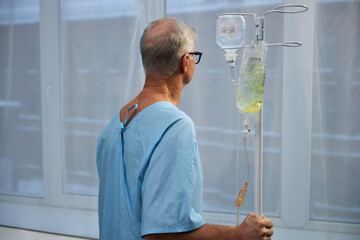Elderly man holding an IV drip while wearing a hospital gown and standing by a window, partially...