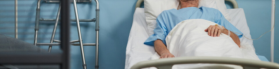 Elderly patient lying on hospital bed covered with blue and white sheets, hooked up to medical...