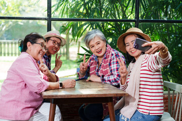 Senior Asian female friends taking selfie cafe smiling cheerful casual clothing sitting together indoor greenery background bright natural light enjoying vacation lifestyle friendship leisure bonding