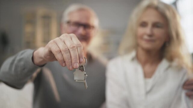 Buy new apartment, keys in hands of elderly man, blurred portrait of aged couple . Mature spouses rejoicing by finish of mortgage, happy house owners, real estate agency, rent, purchase or sale flat