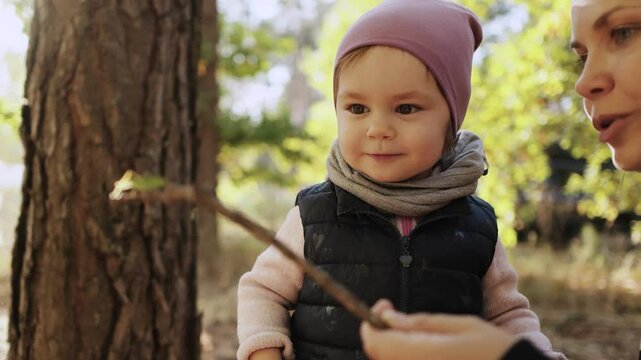 A child curiously looks at a grasshopper on a stick held by her mother, exploring the autumn forest together. Family concept.