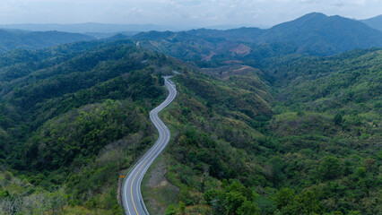 A winding road curves through lush green hills under a cloudy sky, surrounded by dense forests and...