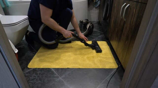 Young cleaner man with vacuum cleaner in hands vacuums shaggy rug in bathroom. Apartment cleaning. Slow motion.